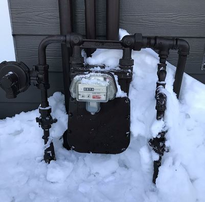 A gas meter and pipes are partially covered in snow outside a building with gray siding. Snow is piled up around the base of the meter and pipes.