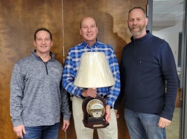 Three men stand indoors, smiling at the camera. The man in the center, wearing a blue plaid shirt, is holding a lamp with a clock at its base. The other two men stand on either side, both wearing casual sweaters.