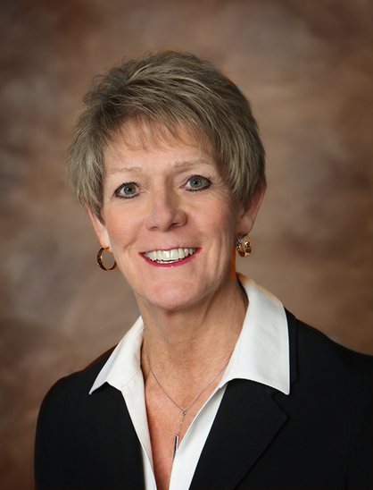 A woman with short light brown hair wearing a dark blazer, white collared shirt, hoop earrings, and a necklace, smiling in front of a brown, softly blurred background.