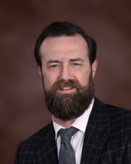 A man with a full brown beard and mustache, wearing a dark plaid suit jacket, white dress shirt, and gray tie, smiles at the camera with a blurred brown background.